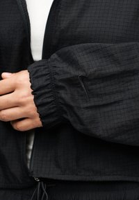 Άτομο που φοράει μαύρο υφασμάτινο μπουφάν με ελαστική μανσέτα και μικρό μαύρο λογότυπο Nike στο μανίκι, πάνω από λευκό ριμπ μπλουζάκι.