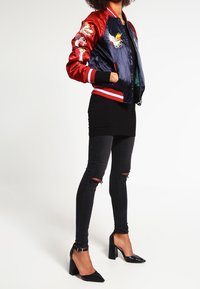 Donna che indossa una giacca bomber ricamata rossa e blu navy, jeans skinny neri strappati e scarpe con tacco alto e cinghietto alla caviglia nere, in piedi.
