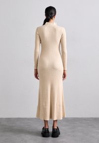 Abito lungo attillato in maglia a coste beige con colletto arricciato e fondo svasato. Abbinato a scarpe platform nere e spesse.