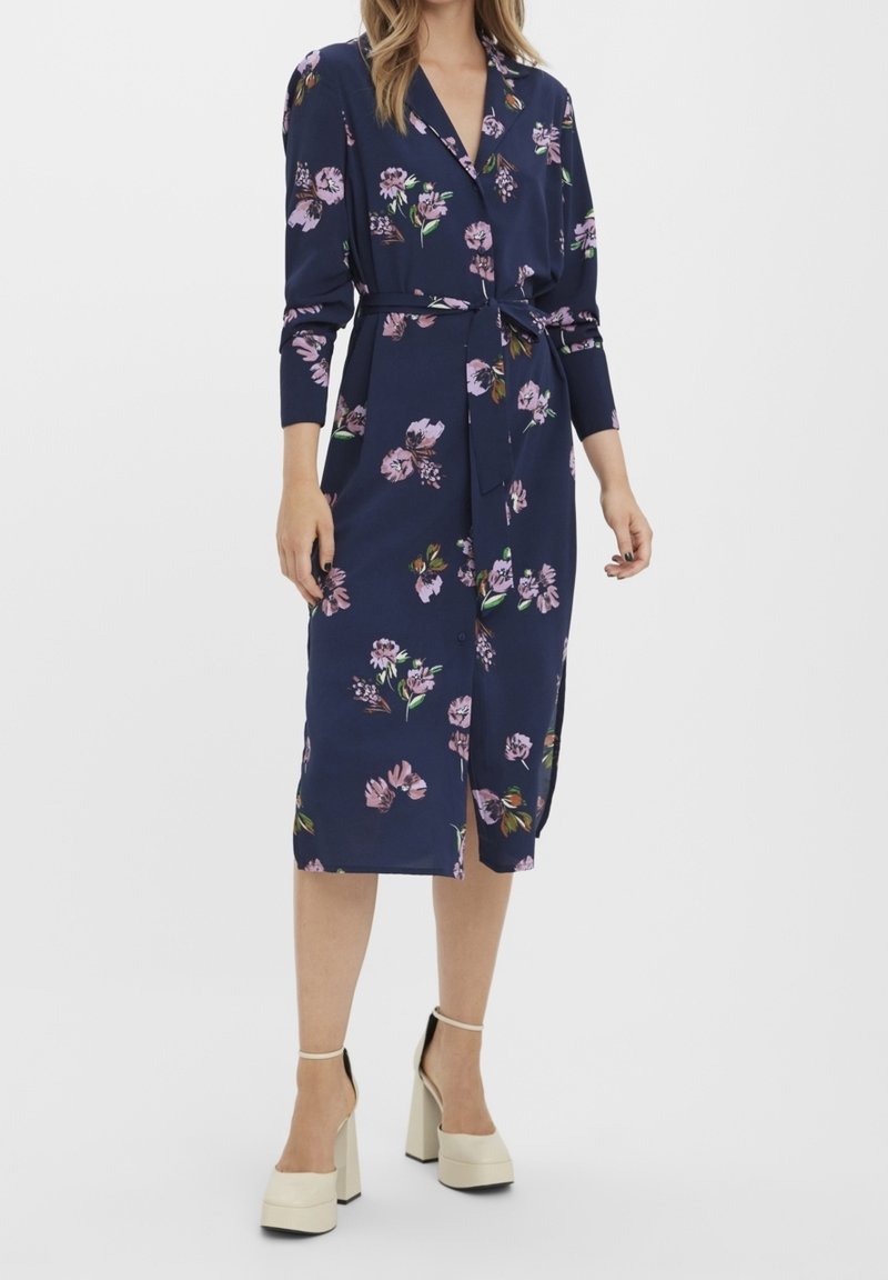 Žena v tmavě modrých květinových midi šatech s dlouhým rukávem a zavazovacím páskem, doplněná béžovými platformovými podpatky.