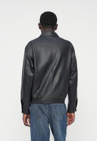 Veste en cuir noir avec col à revers, ourlet élastique et coutures visibles. Le dos présente une texture lisse et une coupe décontractée.