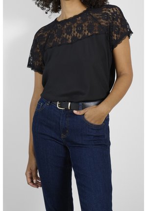 Kvinde iført gennemsigtig sort bluse med blomsterblonde og korte ærmer, stoppet ind i mørkeblå jeans med sort bælte, den ene hånd i lommen.