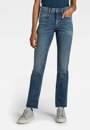 Jeans en denim bleu avec une coupe droite, décoloration légère, cinq poches et un bouton à fermeture, associés à des baskets grises.