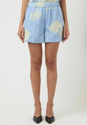 Hele sinised lühikesed püksid beeži lillemustriga, mida kannab seisev inimene paljaste jalgade ja mustade teravate kandadega kingadega.