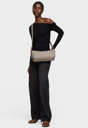 Mujer con blusa negra sin hombros, vaqueros negros de pierna ancha, sandalias de tacón, joyería dorada y llevando un bolso de hombro beige con textura.