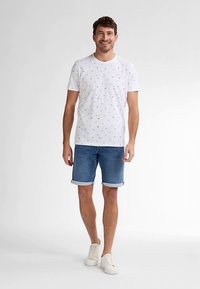 Bílá košile s krátkými rukávy a malými rozptýlenými námořnickými vzory, v kombinaci se středně modrými džínovými šortkami s ohrnutými lemy a bílými teniskami.