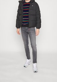 Mustat puhalla takki hupulla, päällä monivärinen raidallinen neule, yhdistettynä harmaisiin slim-fit farkkuihin ja valkoisiin lenkkareihin.