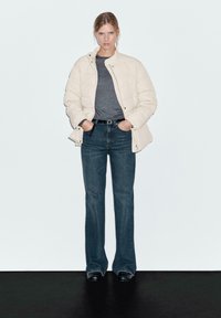 Une femme se tient contre un mur uni, portant une veste matelassée crème, un t-shirt gris foncé, un jean bleu foncé taille haute, une ceinture noire et des chaussures noires.