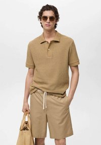 Polo shirt sabbia a coste con colletto e due bottoni, abbinato a pantaloni corti coordinati e una borsa a tracolla intrecciata. Gli occhiali da sole completano l'outfit.