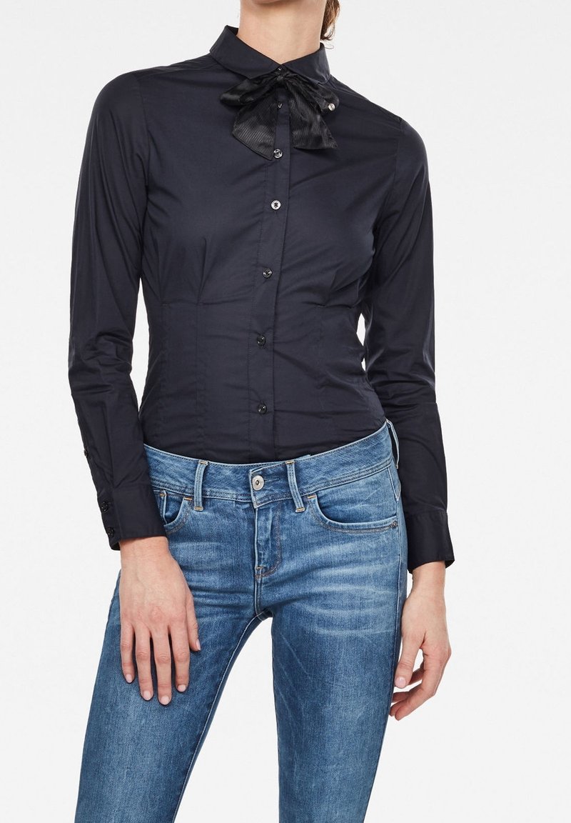 Femme portant une chemise noire cintrée boutonnée avec un col à nœud papillon et un jean bleu en denim, debout devant un fond blanc.