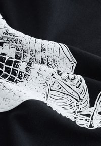 Fekete szövet, rajta merész fehér grafikai nyomat részletes csontvázmintákkal és texturált elemekkel, egyedi mintát mutatva.