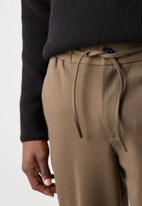 Pantalons de jogging khaki avec une taille à cordon de serrage, présentant une texture lisse et des manches de pull côtelées noires contrastantes.
