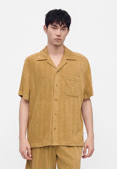 Jeune homme portant une chemise jaune moutarde texturée à manches courtes avec poche poitrine et pantalon assorti, debout devant un fond uni.
