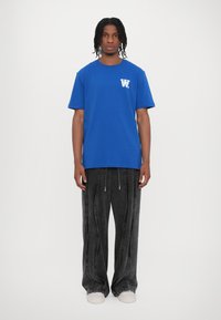 Blue cotton t-shirt with white logo on chest, paired with gray wide-leg denim pants and white shoes, standing on a plain background.