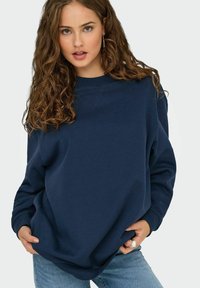 Tamno plavi oversized dukserica izrađena od mekanog materijala, s rebrastim manžetama i rubom, klasičnim okruglim izrezom i opuštenim krojem.