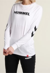 Witte longsleeve shirt met het "HUMMEL" logo in het zwart. Heeft zwarte chevronaccenten op de mouwen en is gecombineerd met zwarte broeken. Eenvoudig ontwerp.