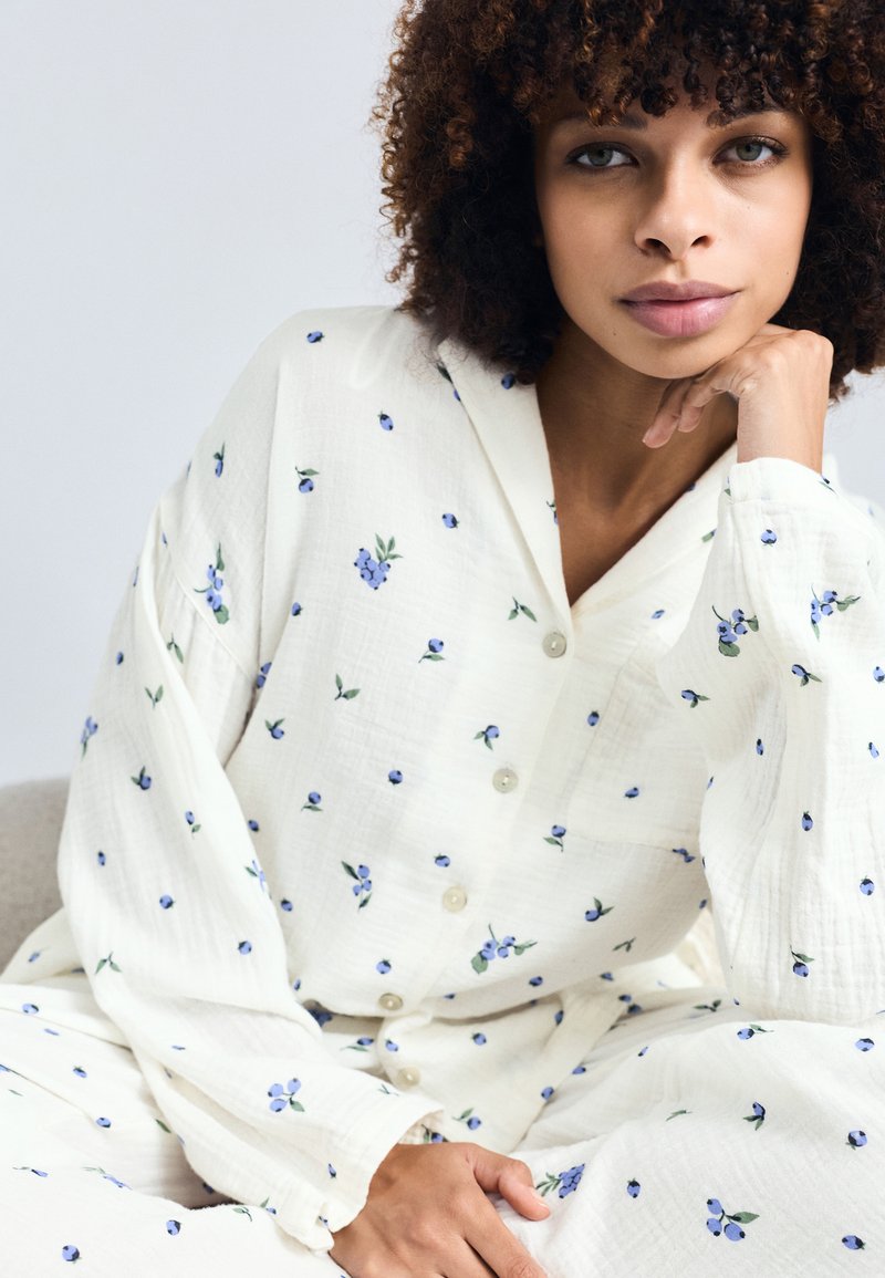 Biała koszula z guzikami, z niebieskimi wzorami kwiatowymi, długością rękawów do dłoni i luźnym krojem. Posiada kołnierz i miękką, teksturowaną tkaninę.