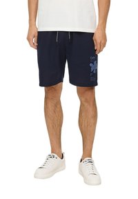 Shorts texturés bleu marine avec cordon de serrage, arborant un imprimé graphique bleu clair sur la jambe inférieure et des poches latérales. Portés avec des chaussures blanches.