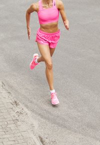 Top corto deportivo rosa y pantalones cortos, corriendo sobre una superficie gris. Zapatillas de deporte rosa brillante con suelas blancas, calcetines blancos visibles.