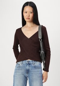 Top marron à manches longues, décolleté en V profond et ourlet droit. Associé à un jean en denim bleu clair et un sac noir avec une bandoulière argentée.