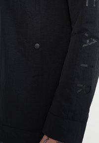 Černá vnější bunda s texturovaným povrchem. Obsahuje zapínání na knoflíky a tištěné detaily na rukávu. Přiléhavý střih a hladká tkanina.