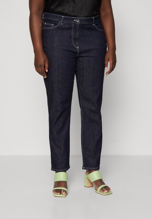 Persoon draagt donkerblauwe rechte jeans en lichtgroene open-toe sandalen met blokhak, staand tegen een effen achtergrond.