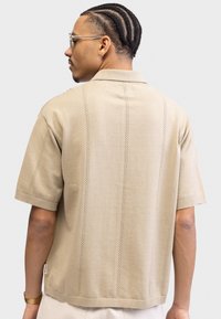 Beige strikket polo-skjorte med korte ermer, har vertikal struktur og liten side-lapp. Glatt tekstur, avslappet passform, minimalistisk design.