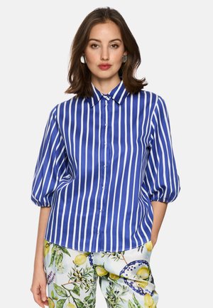 Junge Frau trägt eine blau-weiß vertikal gestreifte Bluse mit Puffärmeln und Blumen-Zitronenmuster-Hose, steht mit einer Hand in der Tasche.