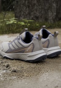 Ζευγάρι γκρι και μπεζ παπούτσια πεζοπορίας Adidas Terrex τοποθετημένα σε μια υγρή, λασπώδη εξωτερική διαδρομή με φόντο γρασίδι και φλοιό δέντρου.