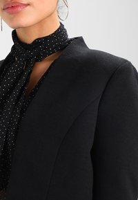 Nainen, jolla on päällään musta bleiseri ja musta pisteellinen huivi sidottuna kaulan ympärille, suurilla hopeanvärisillä renkaankorviksilla, harmaata taustaa vasten.