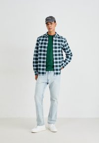 Overhemd met knopen in een groen, marineblauwe en witte ruit; effen groen T-shirt met ronde hals eronder; lichtblauwe jeans; witte sneakers; grijze pet.
