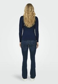 Top de manga larga acanalado de color azul marino combinado con jeans acampanados de lavado oscuro y zapatillas blancas; mostrado desde la espalda con cabello largo y ondulado rubio.