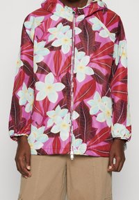 Floral jacket in lightweight synthetic material, featuring large white and blue flowers on a pink background with maroon leaves. Hooded design.