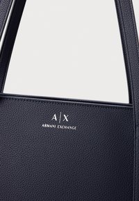 Mornarska kožna torba s teksturiranom površinom, čvrstim trakama i utisnutim logom "A/X ARMANI EXCHANGE" u bijeloj boji.