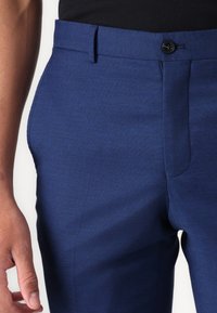Pantalon bleu marine avec une texture lisse, coupe ajustée, fermeture à bouton et poches inclinées. Détails de coutures visibles sur le côté.
