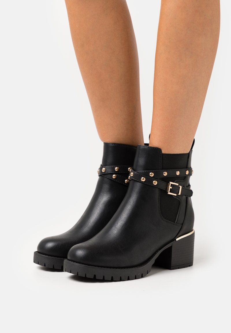 Bottes noires en cuir à talons épais, avec des sangles cloutées et des panneaux élastiques latéraux, portées sur des jambes nues sur un fond uni.
