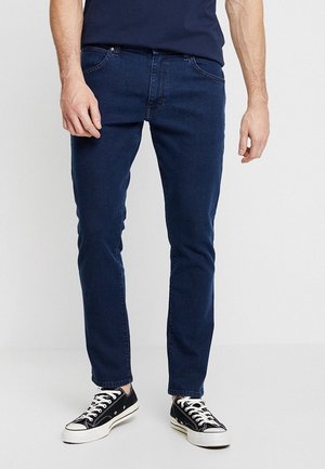 Mężczyzna w ciemnoniebieskich obcisłych dżinsach, czarno-białych płóciennych trampkach i granatowej koszulce, stojący na jednolitym tle.