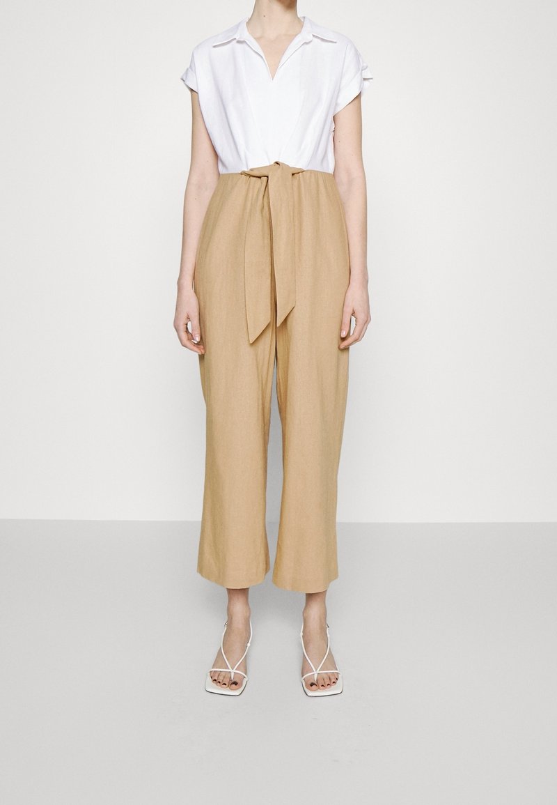 Personne portant un pantalon beige taille haute avec un nœud sur le devant, un chemisier blanc à manches courtes et des sandales blanches à talons avec des lanières.