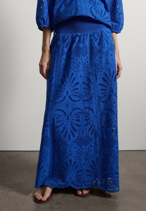 Vrouw draagt een lange blauwe eyelet kanten rok en een bijpassend topje, staat blootsvoets op een grijze vloer met bruine sandalen.