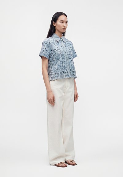 Femme debout portant un pantalon large blanc, une chemise en dentelle bleu clair à manches courtes et des sandales marron sur un fond blanc uni.
