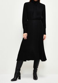 Blouse noire à manches longues avec un col haut et un détail de nœud, associée à une jupe noire au genou et des bottes noires à talons hauteur genou.