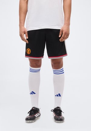 Nogi w czarnych szortach piłkarskich Manchester United, białe skarpety za kolano z niebieskimi paskami i czarne korki Adidas na białym tle.