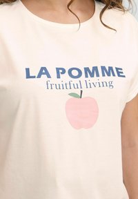 Šviesiai kreminių spalvų medvilnės marškinėliai su trumpomis rankovėmis, ant kurių yra mėlynas užrašas "LA POMME" ir rožinė obuolio grafika su žalia lapo detalė.