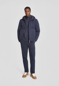Giacca a vento blu navy con cappuccio, tessuto strutturato, chiusura con zip, indossata sopra una maglia a maniche lunghe scura, abbinata a pantaloni blu navy e sneakers marroni.