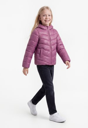 Jong meisje loopt, draagt een roze donsjas, zwarte broek en witte sneakers, glimlachend tegen een effen achtergrond.
