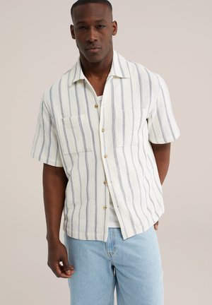 Mężczyzna ubrany w białą, krótkorękawową koszulę na guziki z pionowymi niebieskimi paskami oraz jasnoniebieskie dżinsy, stojący na jednolitym tle.