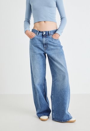 Person iført lyseblå cropped langærmet top, løstsiddende blå jeans med høj talje og hvide sneakers, hænder i lommerne, stående på hvidt gulv.
