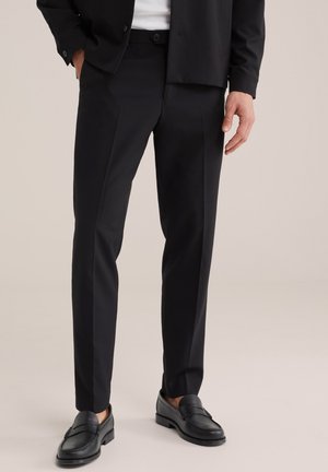 Mand iført sort, tætsiddende bukser, sorte loafers og en delvist synlig sort jakke over en hvid skjorte, stående med den ene hånd i lommen.