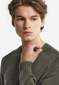 Olivgrön tröja med ribbat rundhals, texturerat tyg och åtsittande ärmar. Modellen rör vid sitt nyckelben för att visa detaljer.