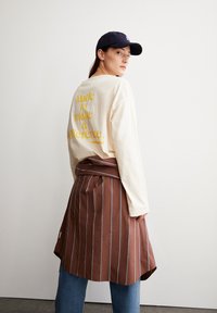 Een crèmekleurig shirt met lange mouwen en gele tekst, gelaagd over een bruine gestreepte wikkelrok. Een donkere pet maakt de outfit compleet.